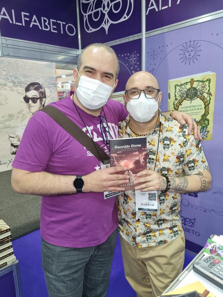 Milo Ambrus e Claudiney Prieto dando um abraço, segurando o livro Escuridão Eterna: A Fúria dos Anjos.